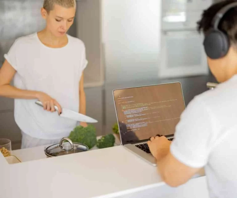 A woman cooking brocolli while a man works on a laptop.
